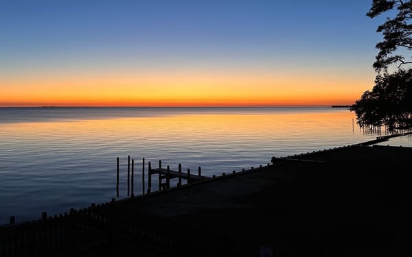 Early morning. Sunrise colors in the sky, which is reflected in the almost still waters of the Roanoke sound. There's a short pier visible in silhouette, either in ruins or half-constructed.
