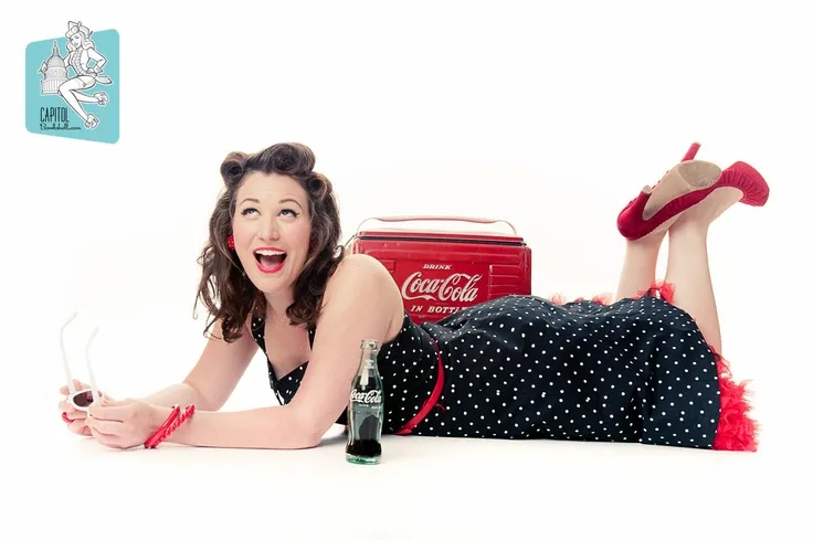 A woman laying on her stomach, wearing a polka-dot dress, smiling. There is a Coca-cola branded cooler and a Coke bottle nearby.