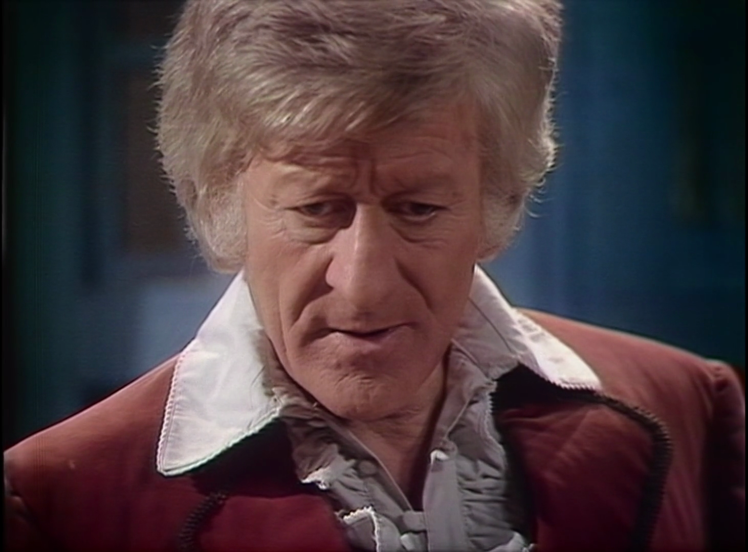 Jon Pertwee as the Doctor, wearing a red jacket and a frilly light-colored shirt with huge 70s collars.